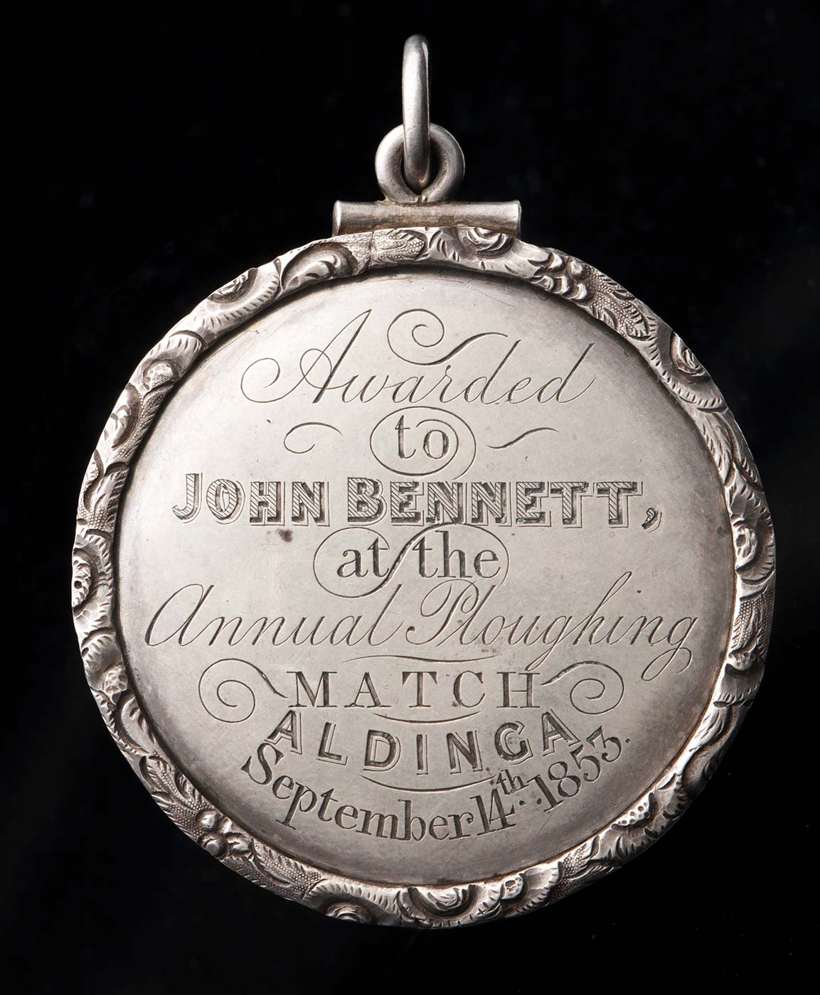 A silver coloured agricultural medal. Engraved text reads 'Awarded / to / JOHN BENNETT, / at the / annual ploughing / MATCH / ALDINGA / September 14th 1853. - click to view larger image