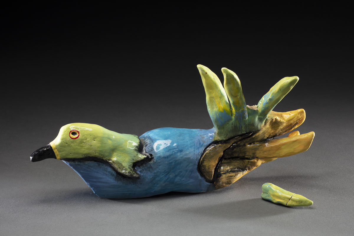 A small culpture of a bird with a green head, blue body, green and yellow tail feathers. One of the tail feathers has broken off and sits beside the bird's body.