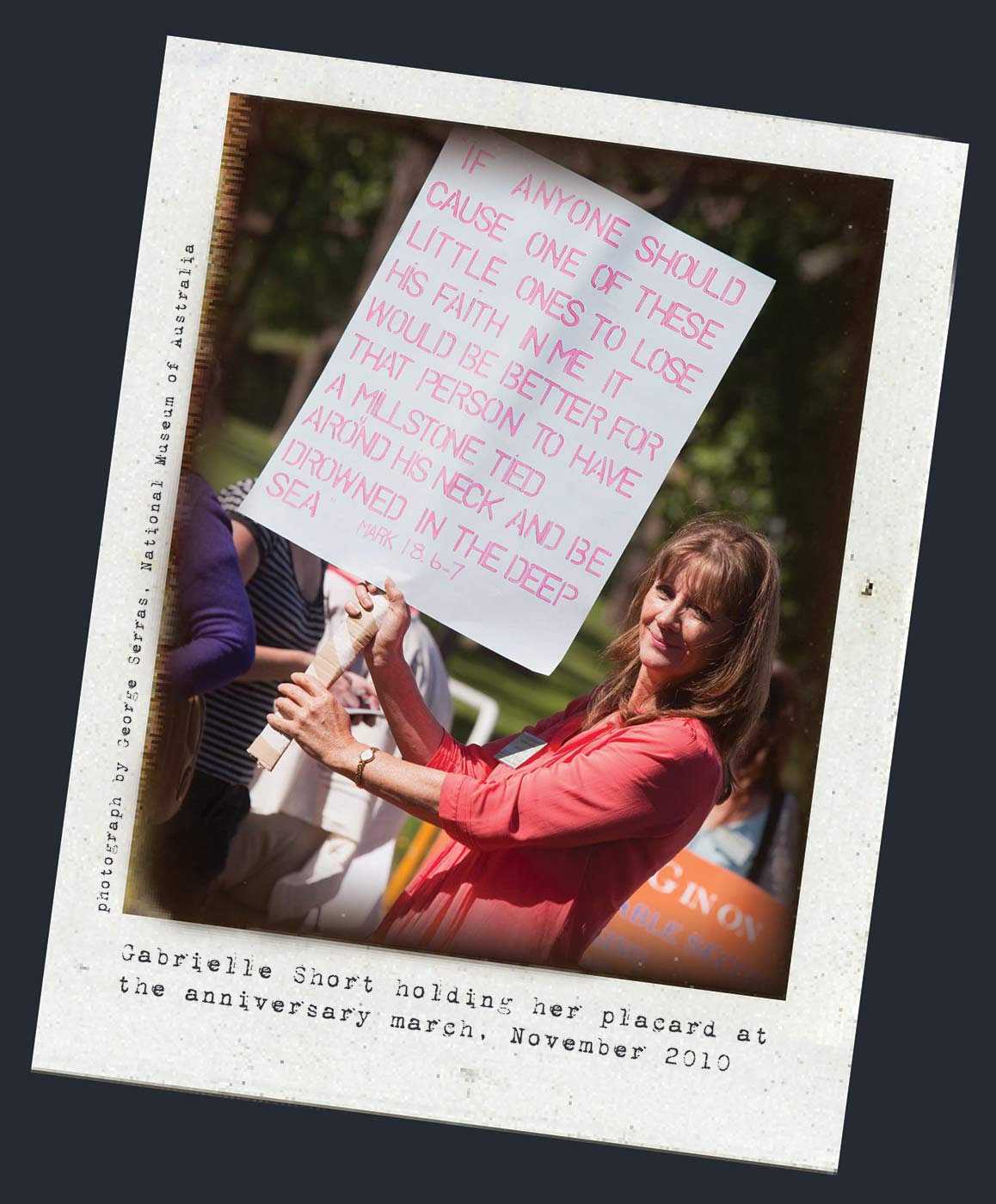 A Polaroid photo of a woman holding a banner on a white cardboard sheet. Typewritten text below reads 'Gabrielle Short holding her placard at the anniversary march, November 2010.' 'Photograph by George Serras, National Museum of Australia' is printed along the left side. - click to view larger image