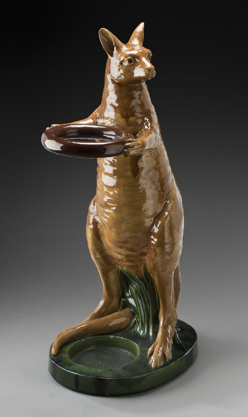 Ochre-glazed stoneware kangaroo, standing on a base. The kangaroo's paws extend around a ring and its feet and tail straddle a circular recess in the base. - click to view larger image