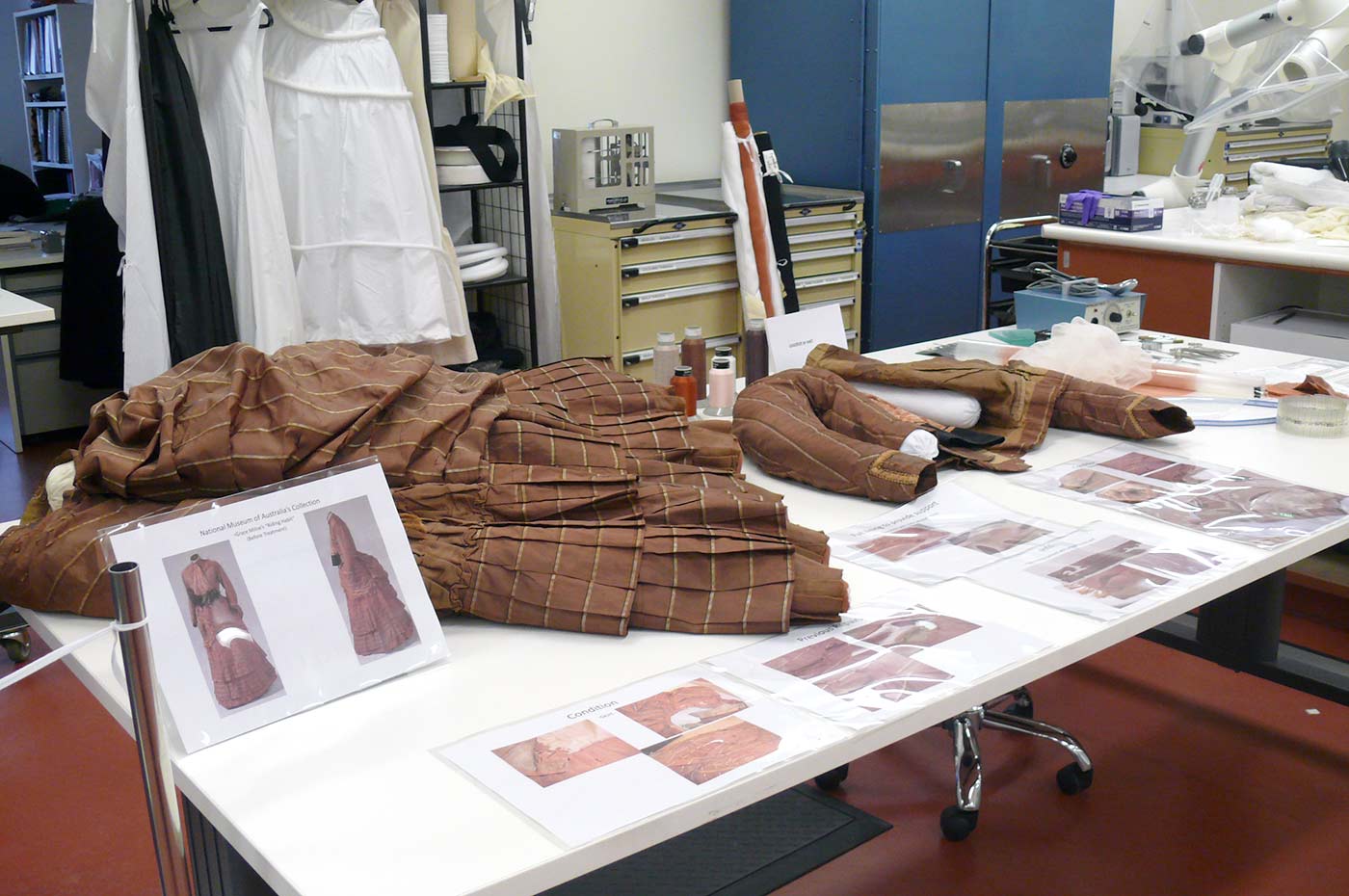 Colour photograph showing a full, brown skirt and small jacket laid out on a table in a laboratory. Images of the complete outfit and detailed sections are laid on the table too. - click to view larger image