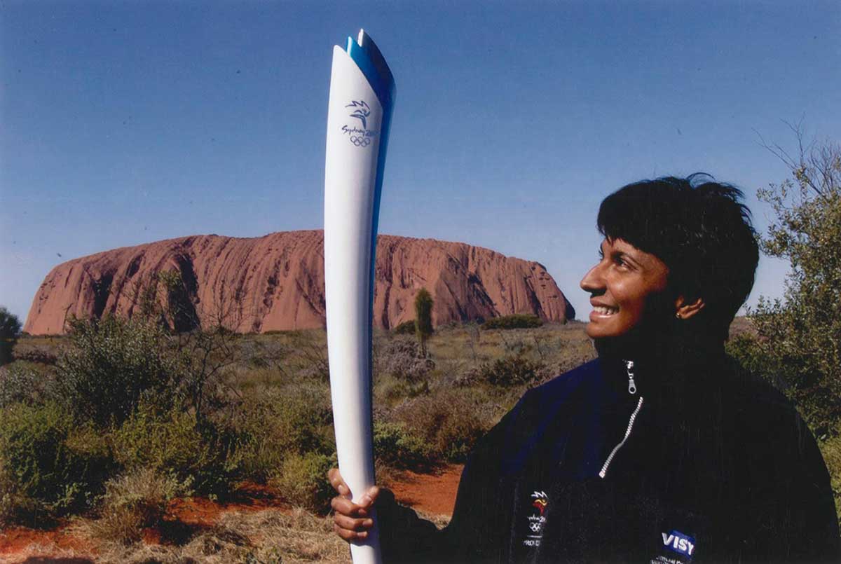 Colour photo of a woman holding up the Sydney 2000 Olympics torch with Uluru in the background. - click to view larger image