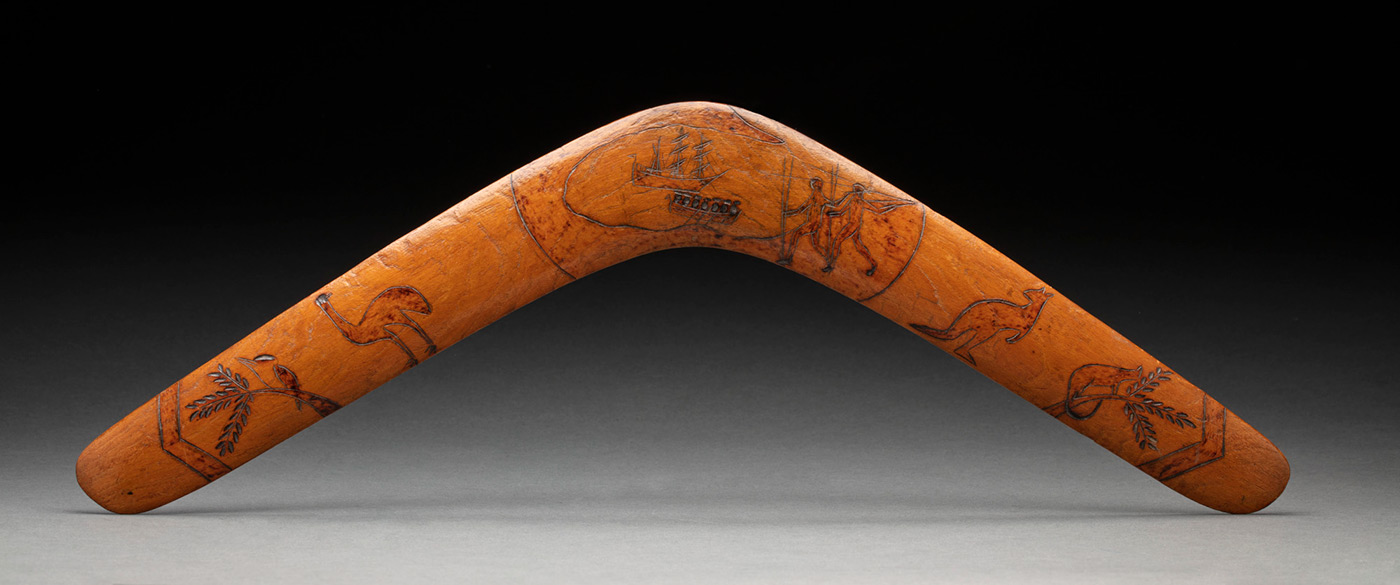 Wooden boomerang featuring illustrations incised onto the surface. The illustrations feature images of plants and animals, a ship with a rowboat nearby and people with spears.