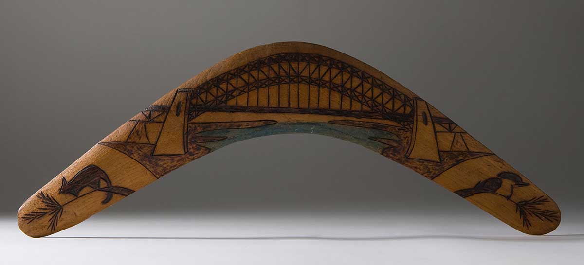 Boomerang made of light-coloured wood, depicting the Sydney Harbour Bridge in the centre, with a possum to its left and a kookaburra to its right.