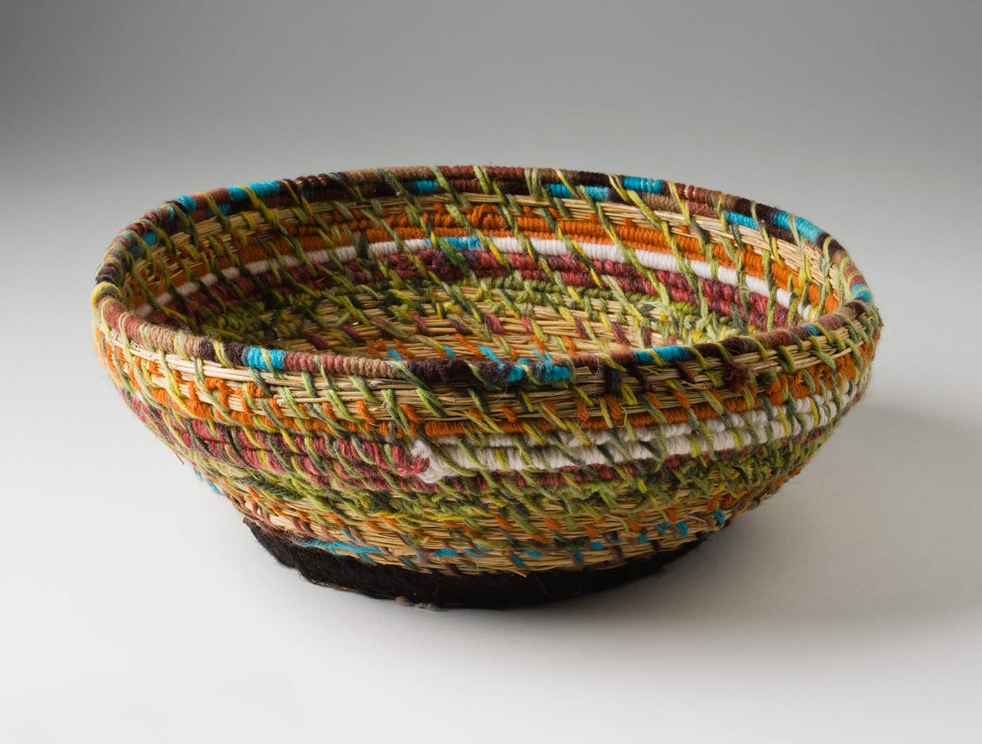 A circular coiled yarn and plant fibre basket with a flat metal base. The base is made of a blue 'Fray Bento / Steak and Kidney Pie' tin lid which has been attached to the yarn section with black yarn through holes punched in the metal. The coiled yarn in muted tones of green, orange, yellow,pink, turquoise, white and orange, is spaced out so the plant fibre centre can be seen. - click to view larger image