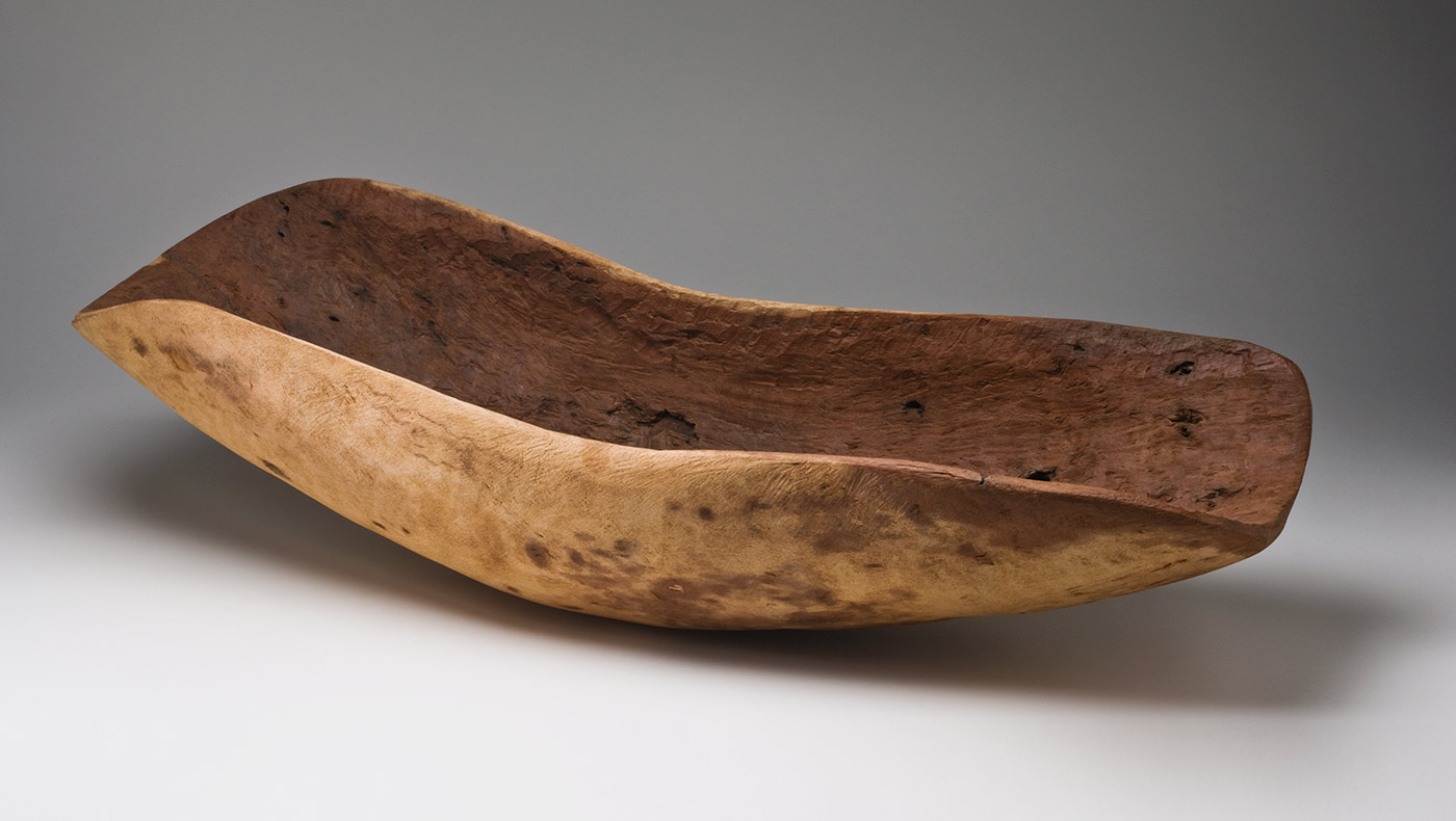 A beige brown wooden concave oval container with ends that curve up. The outer surface is smoothed and shows the grain of the wood, as well as some darker stain-like patches. The inner surface is a red-brown colour and has a rough texture with unevenly shaped holes.