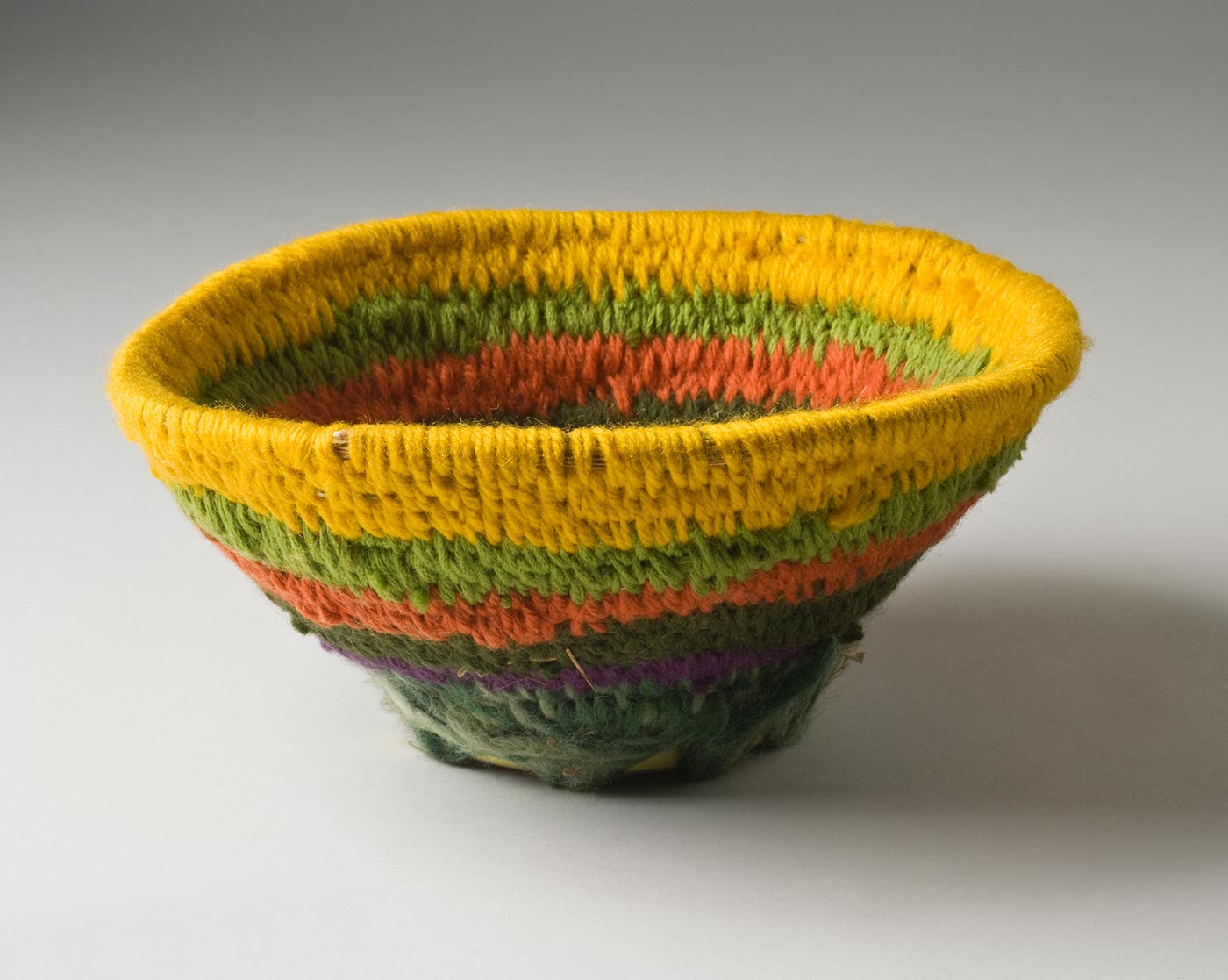 A circular coiled striped yarn basket over plant fibre, with a metal base. The basket has a circular tobacco tin base with text on the back and holes punched around the base. The yarn coiled fibre section is attached with yarn through the holes in the tin, and opens out toward the top. The basket has a stripe of yellow at the top edge followed by green, orange, dark green, purple and white-green. - click to view larger image