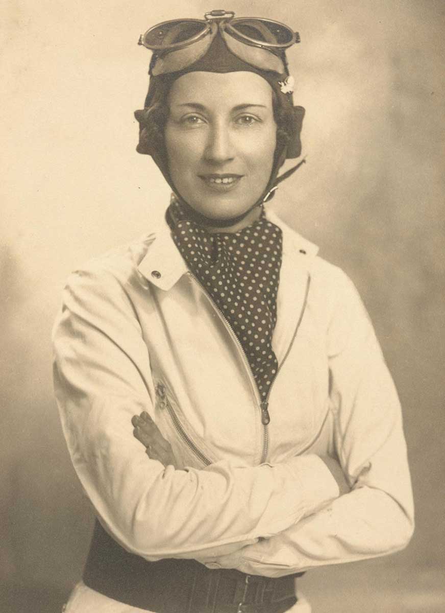 Black and white portrait photograph showing Joan Richmond standing with her arms crossed. She is wearing a white racing jacket with a black and white polka dot scarf. On her head is a racing cap and a pair of goggles. - click to view larger image