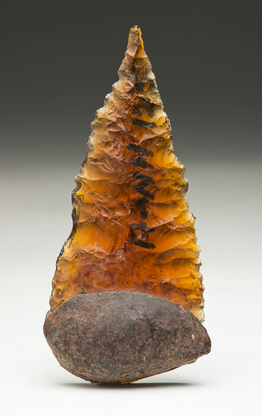 Clear brown glass Kimberley point or spearhead. It is bifacially flaked with serrated edges, rounded at the proximal end, and tapers to a sharp point. A knob of adhesive is attached to the rounded end on one surface only. - click to view larger image