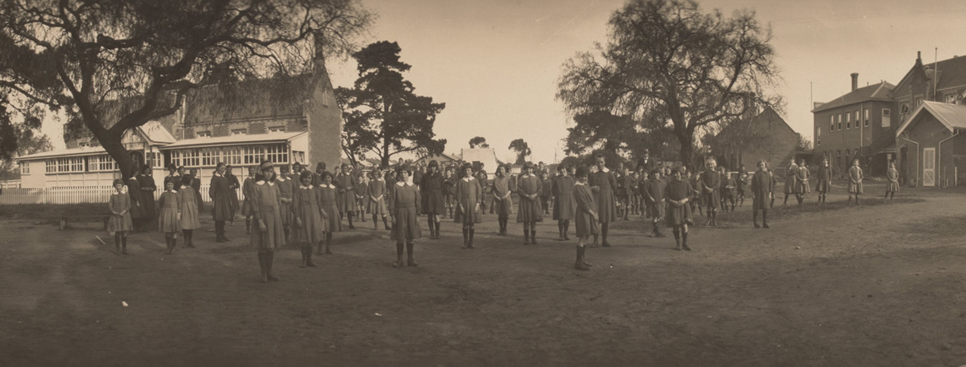 Cropped black and white photograph of a large group of children standing in the grounds of an institution.