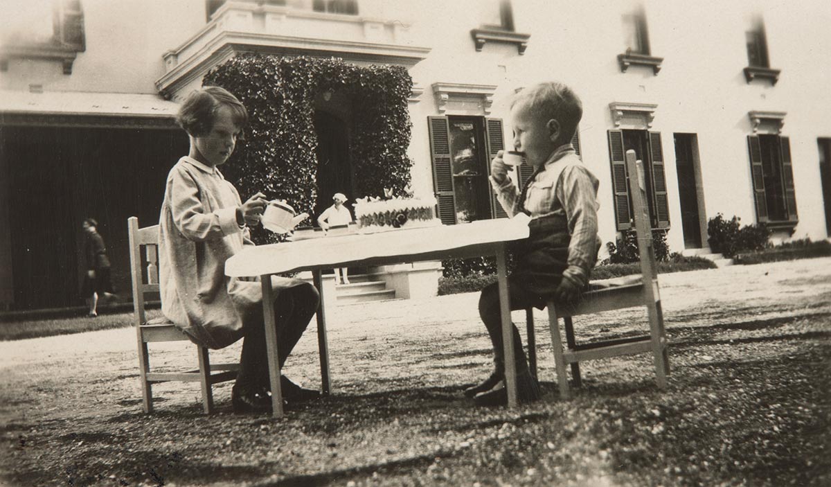 Black and white photograph of two children seated at a low table in front of a house. - click to view larger image