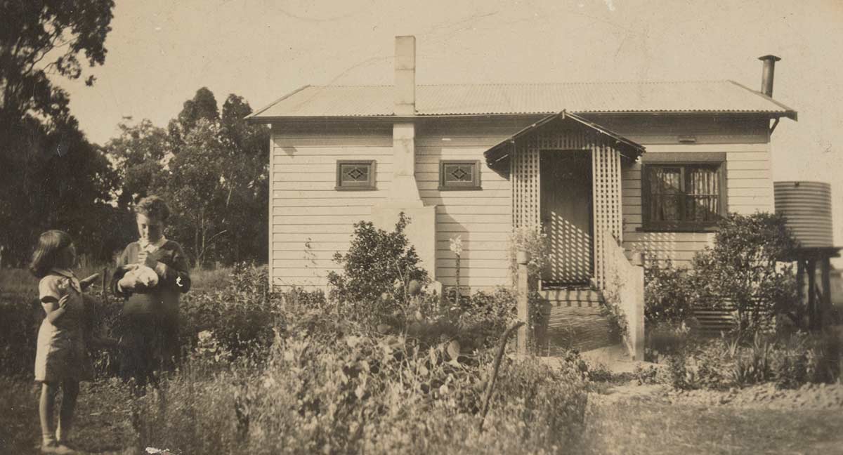 A black and white photograph of a young girl and boy standing in a garden in front of a weatherboard cottage.  - click to view larger image