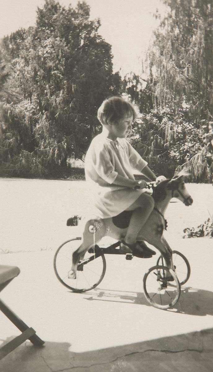 Black and white photograph of a young girl seated on a horse-shaped tricycle. - click to view larger image