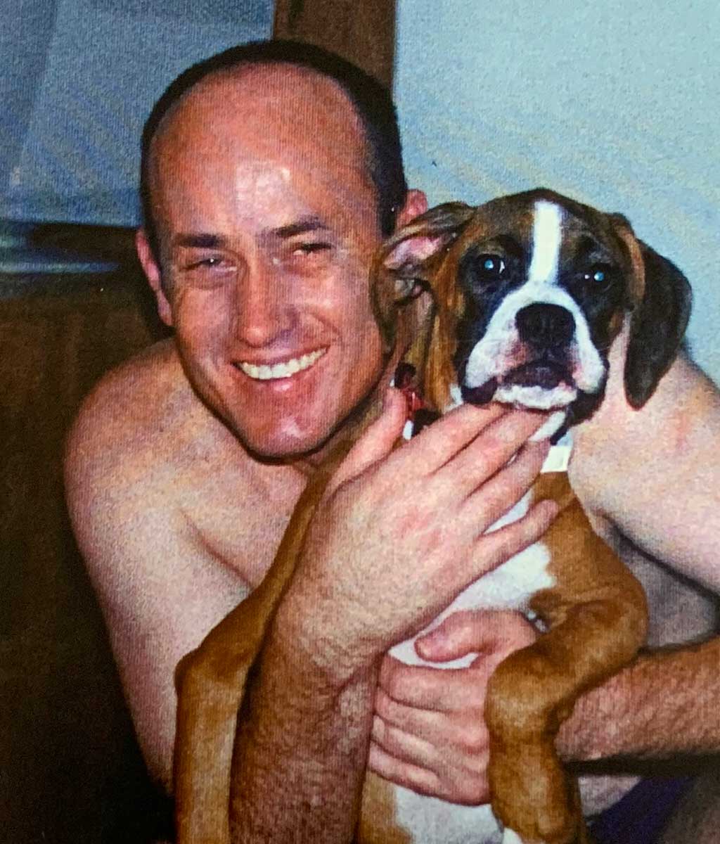 Front view of a shirtless man crouching and holding a boxer puppy in his arms. The man is smiling broadly. - click to view larger image