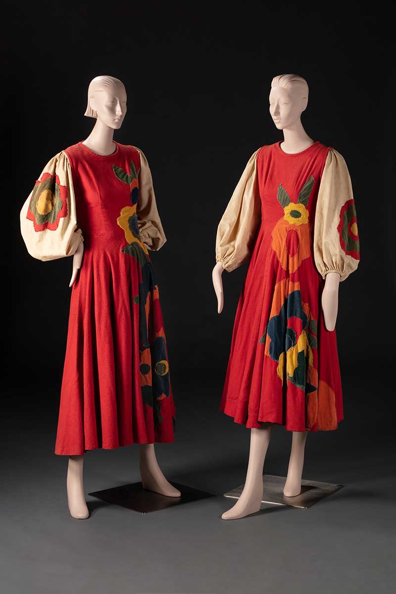 Two mannequins displaying costumes featuring a full length red dress with loose beige sleeves, and floral embellishments. - click to view larger image