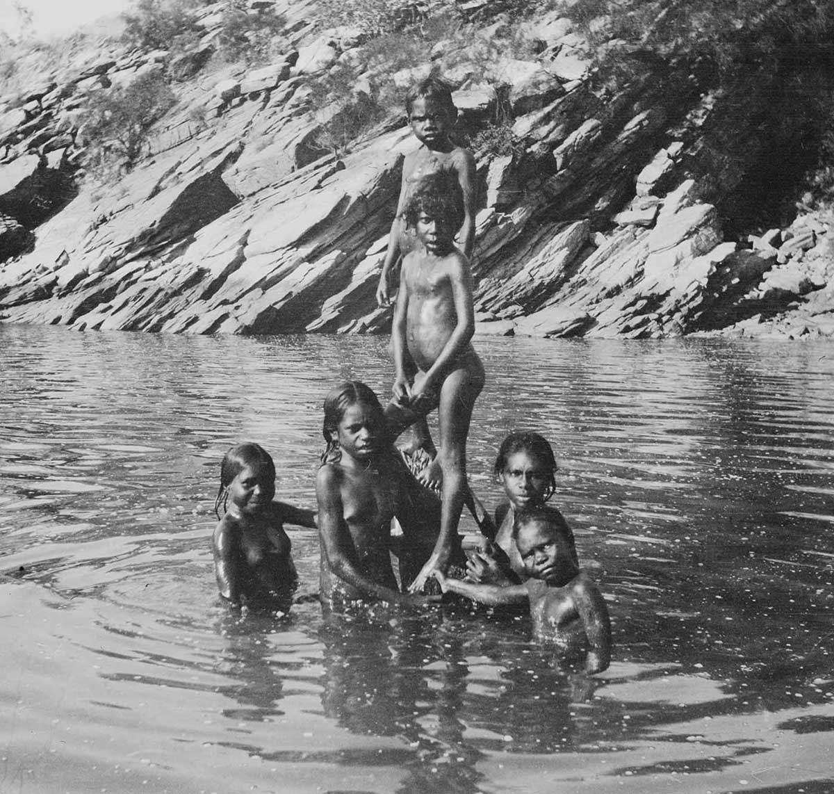 Aboriginal children in a rock pool, Sunday Island, Western Australia, 1916. The six children are on and around a partially submerged mangrove stump. Two of the children stand out of the water on the stump. The rest are visible from about their waists up. The water is rippling in concentric circles around the children. In the background is part of the rock pool bank. It is a rocky slope with large flat weathered sections. Bushes grow in places along the slope. A larger and more dense patch of bush can be seen at the right side of the image. The reflections of the children and bank on the water are broken up by the rippled surface. - click to view larger image