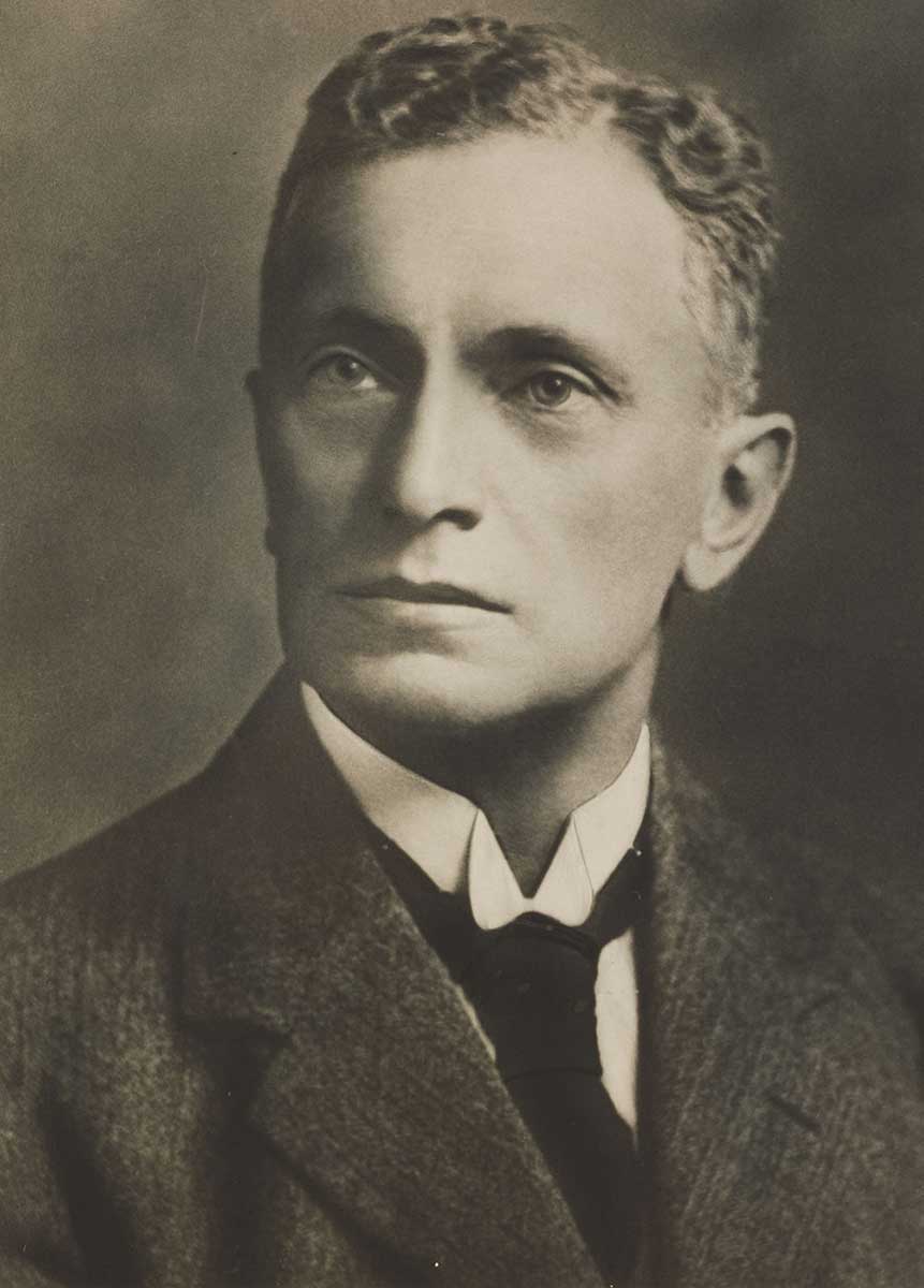 Portrait of Herbert Basedow, about 1920. He is seen from about mid-chest up. His shoulders face to the right of the image while he has his head turned to the left of the image. He wears a jacket, light coloured shirt with high, starched collar, and a dark tie. His short, wavy hair is neatly combed and tidy. His expression is one of earnestness and resolve, as he directs his gaze toward some imaginary landform or horizon. The background is relatively dark and featureless, suggesting a formal studio portrait. The left side of his face, from the viewer's perspective, is in shadow. - click to view larger image