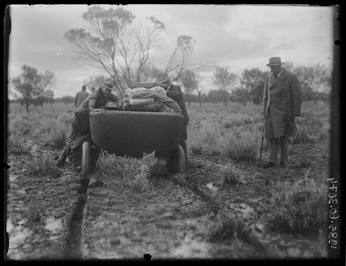 An expedition vehicle bogged on the Burt Plain, Northern Territory 1923. The vehicle, an open-topped car carrying expedition luggage, is bogged on a plain sparsely covered by knee-high bushes and scattered trees. The car is seen from behind; two deep wheel ruts lead from it to the image foreground. Two non-Aboriginal men are attempting to free the car. They are at each side. A third non-Aboriginal man in a suit and long coat stands to the right, watching them. Two other non-Aboriginal men are visible ahead of the car, with their backs to the camera. They appear to be studying the way ahead. The sky is filled with a heavy overcast and the threat of more rain. - click to view larger image