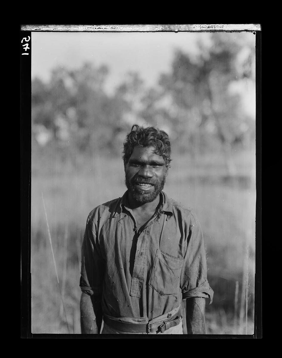 An Aboriginal, Neighbour (or Ngaller-iga), near Roper Bar, Northern Territory 1928. He stands facing the camera, visible from about the waist up. He wears trousers and a long sleeve shirt with the sleeves rolled up to his elbows. His hair is cut in a more European style and he has a full beard. He smiles at the camera in an enthusiastic and friendly manner. Neighbour appears to be relatively young, perhaps in his late twenties. The background of grass and scrub is out of focus in contrast to the sharp and clear image of Neighbour. - click to view larger image