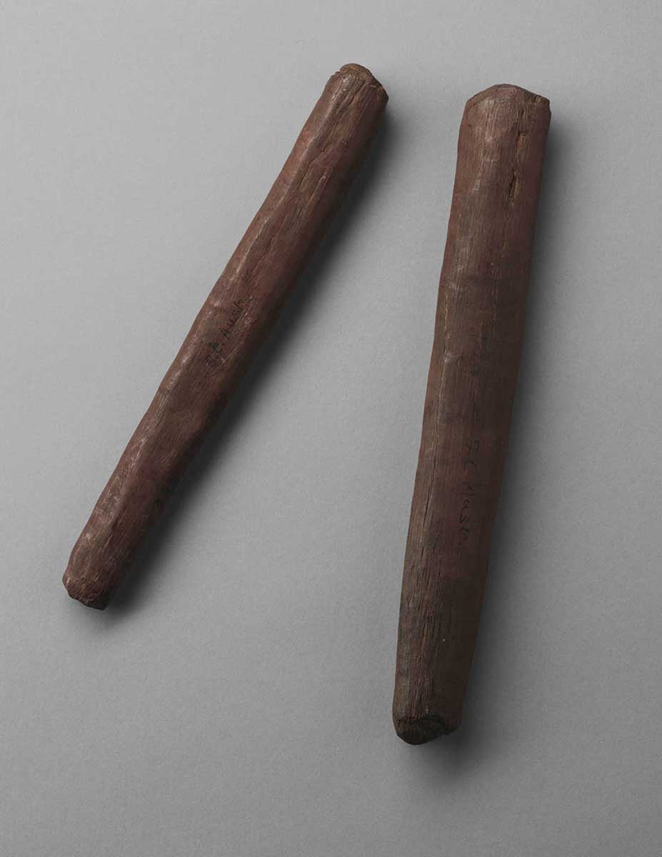 A pair of wooden clap sticks photographed on a grey studio surface. The sticks are about the same length, with roughly rounded-off ends. They are made from hardwood, which has a very close grain. This gives the sticks a hard appearance, as if they were made from metal or iron.  - click to view larger image
