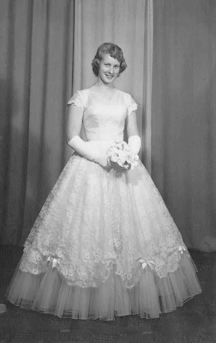 Black and white portrait photograph of a young woman in a long white gown made of a tulle skirt, lace and bow trimmings. She is wearing long white gloves and is holding a small flower bouquet. - click to view larger image