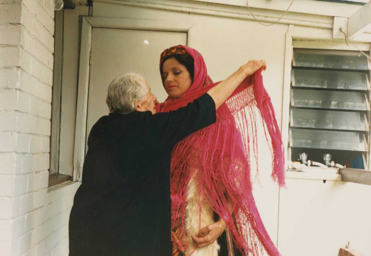 Two women standing facing each other. The woman on the left is older, with short grey hair and wearing a black dress with elbow-length sleeves. She is tying a pink shawl around the neck and shoulders of the younger woman on the right with dark-coloured hair. The woman on the right is wearing a traditional Greek garment, and the pink shawl has long fringed edges which drape to below her hips. The woman on the left has her arm raised as she wraps the shawl around the neck of the younger woman. The two women are looking at each other.