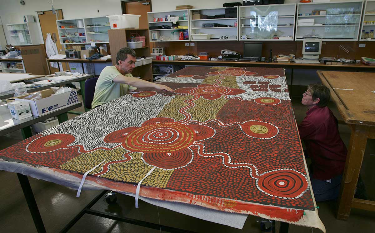 Two conservators work on either side of a large canvas by Uta Uta Tjangala. - click to view larger image