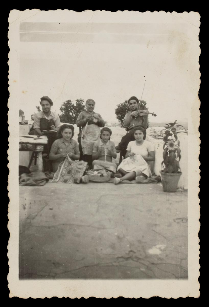 Black and white portrait-oriented photograph of Effie Kyprios and her family. The family sit on a terrace outside their home in Kythera, and each family member holds an item which is special to them. Seated in a chair in the centre is Effie’s mother, holding a tool for spinning yarn. To the left of her is seated Effie’s eldest brother, holding tools. To the right of Effie’s mother is seated Effie’s next brother, holding a violin. Effie and her sisters sit on the ground below them, Effie on the left holding a woven bedspread. Next to her in the middle is Effie’s youngest sister, holding a crochet hook. Effie’s other sister sits to the right, holding an embroidered item. - click to view larger image