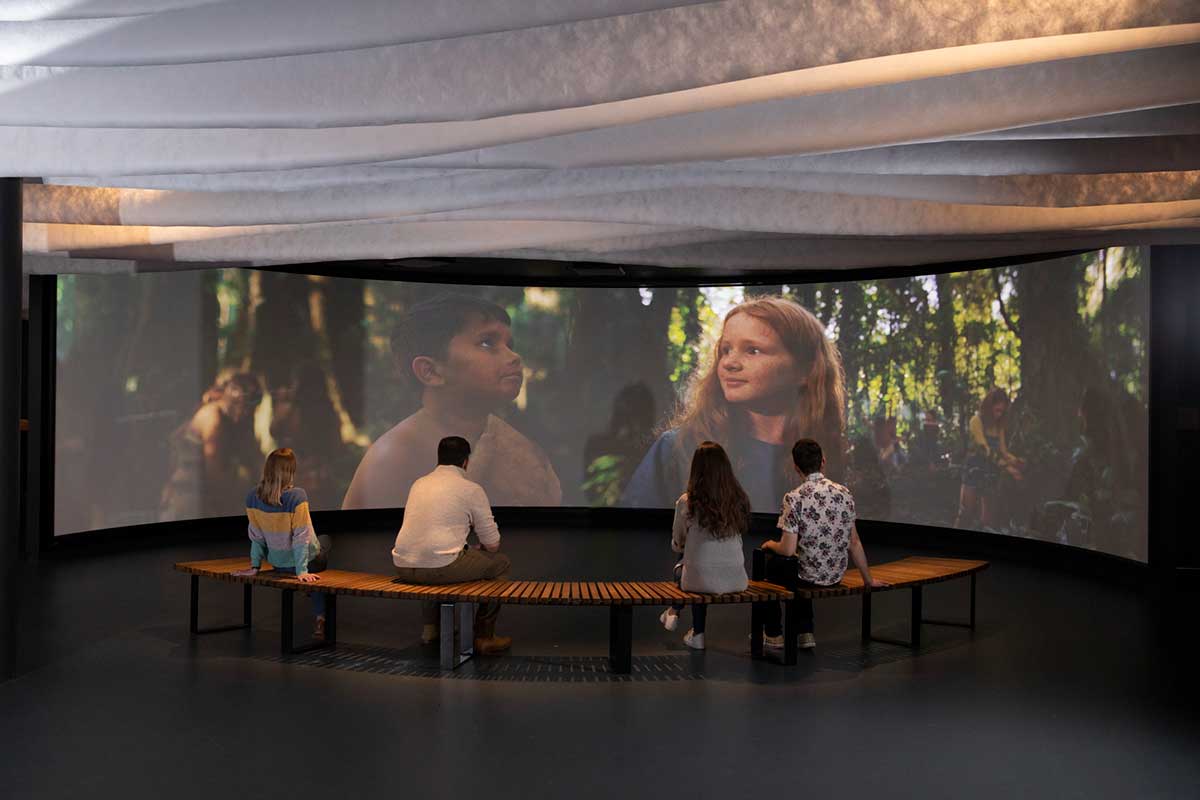 Four people sit with their backs to the camera, facing a large screen which shows two children and other people in a forest setting. - click to view larger image