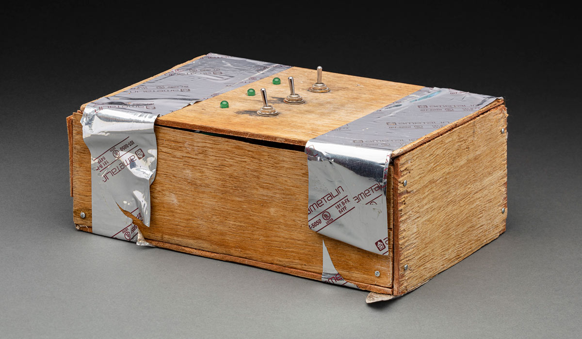 A control panel made from pieces of wood nailed and taped together to form a box. On the top are three silver, metal switches and three green, glass lights. - click to view larger image