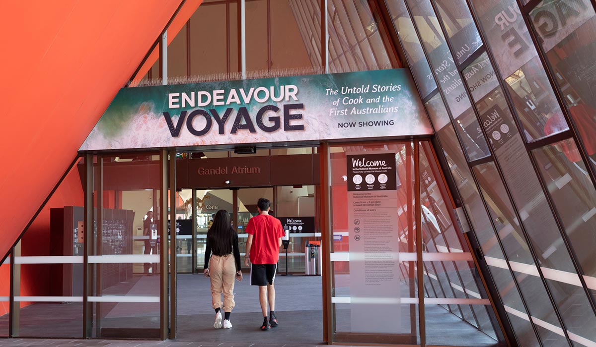 Two visitors walking through a large entrance with a promotional banner for the Endeavour Voyage exhibition above.