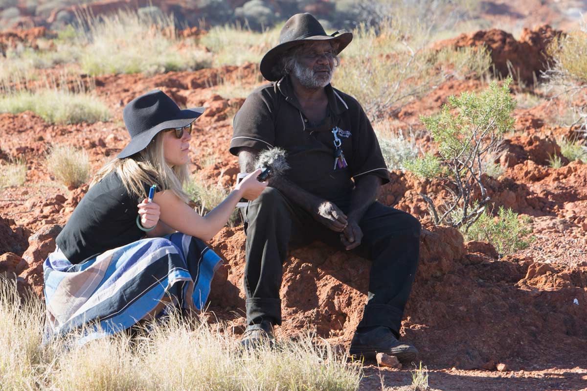A woman records a man as they sit in an outback setting.
