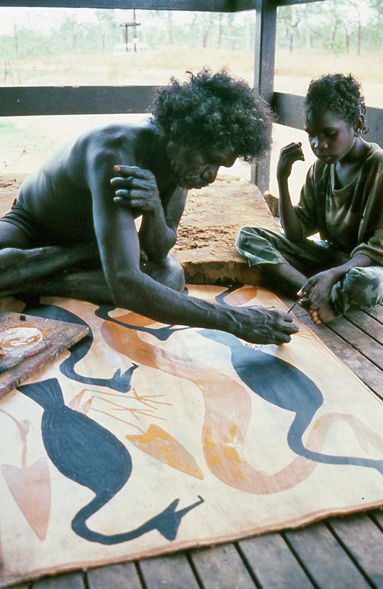 Colour photograph of a man and child sitting on the ground painting. - click to view larger image