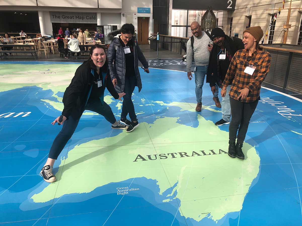 Five people standing on the area of Australia of a large world map printed onto the floor of a public building. - click to view larger image