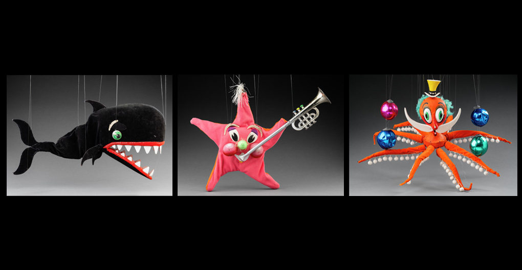 Left: A marionette of a black whale with green eyes. The whale’s mouth has red lips and white triangle shaped teeth. Middle: A marionette of a pink and orange starfish with a green nose and two large red cheeks. At the top of the starfish is a tuft of white ribbons that look like hair, and in its left hand is a silver and gold trumpet. Right: A marionette of an orange octopus and a white moustache wearing a yellow hat. Accompanying the puppet are plastic balls in blue, green and pink.
