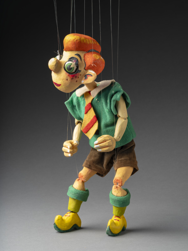 A photograph of Nicky the puppet, he has bright orange hair and is wearing a green short sleeve shirt, brown shorts and a red and yellow tie. On his legs are green socks and yellow clog-like shoes.