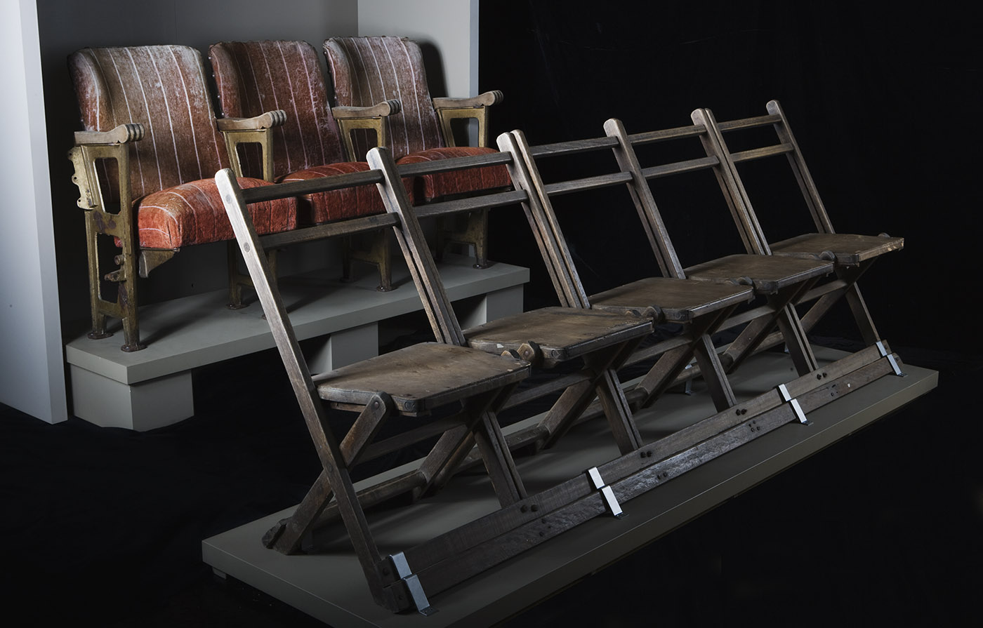 Two rows of theatre seating. Five plain wooden chairs in front of a row of three cushioned armchairs. - click to view larger image