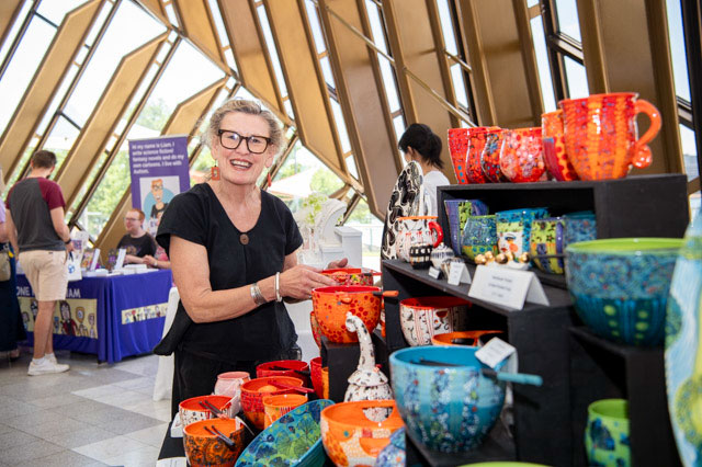 A woman stands holding a bright orange ceramic pot, one of many pots part of a display at a market stall. - click to view larger image
