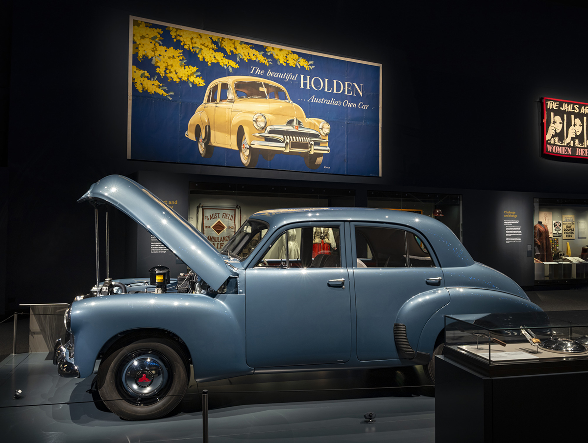 A photograph of a blue Holden car in the Landmarks gallery. Behind the Holden car is a large vintage poster of a Holden.