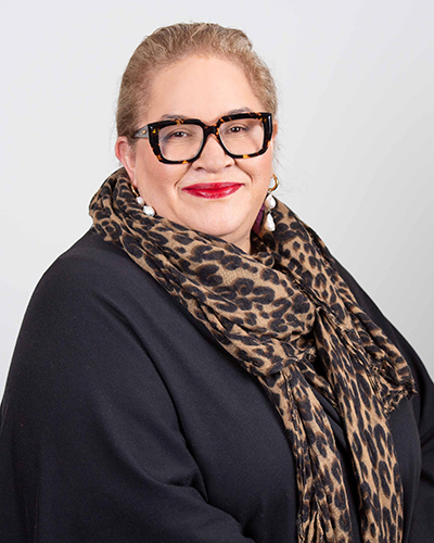 A photograph of Megan Davis. Megan wears a black and orange print scarf, a black shirt and glasses.
