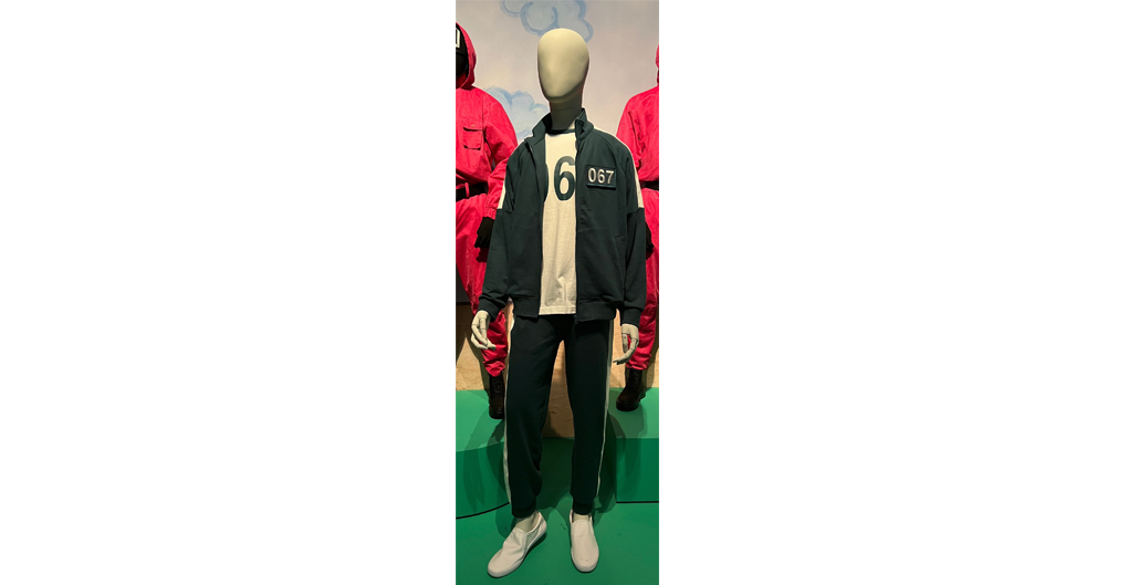 This is a full-size mannequin figure made of fibreglass wearing a green tracksuit and tee shirt, the iconic costume from the Korean series Squid Game. The tracksuit consists of a dark free zip-up jacket, unzipped, and matching tracksuit pants with white side stripes running the length of the arms and the legs. Beneath the tracksuit jacket is a white T-shirt emblazoned with number 6. This number (6) is repeated in a white box on the left side of the jacket. The tracksuit and T-shirt are slightly oversized, paired with plain white sneakers. 