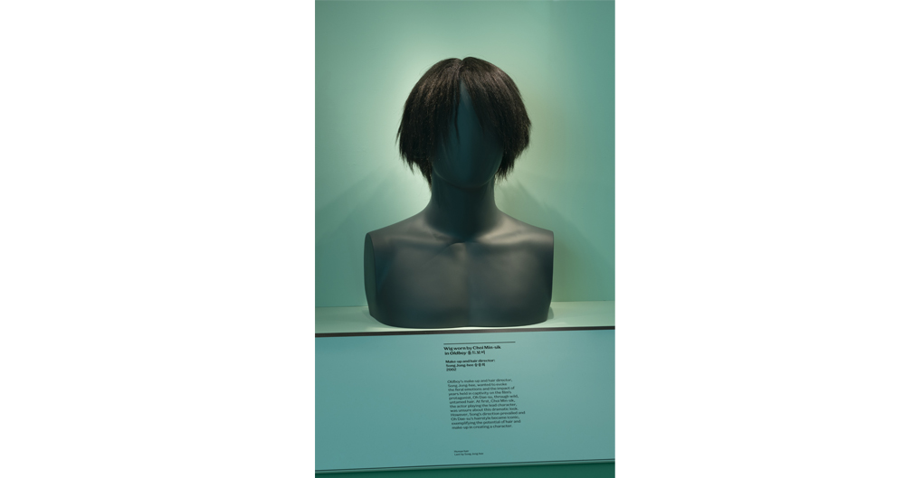 A black torso features a wig on the head. The wig is characterised by a central part and straight, longish hair, which is shaggy and falls below the ears but above the shoulders. The overgrown, unkempt style falls straight over the face and over the ears of the mannequin head.