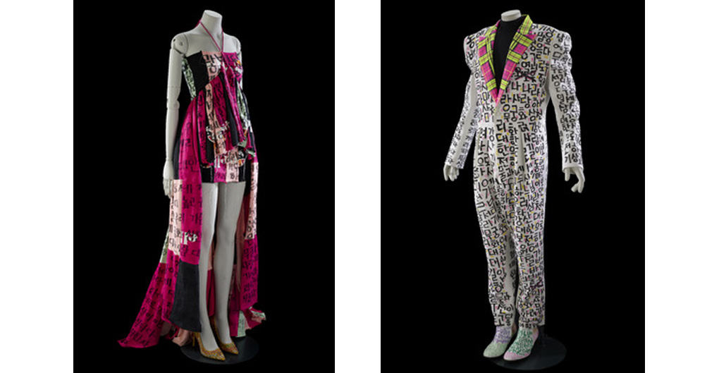 The suit designed by Balko Kim fuses traditional Korean elements with a retro western look. The narrow, well-fitting white suit is covered in black Hangul (Korean alphabet) characters in a hand-drawn style. The lapels feature fluorescent neon pink and green tartan. The shoulders are defined with shoulderpads.  The pants are in an identical white fabric printed with hand painted black hangul.  The narrow ankle boots are rainbow coloured.    The dress is an asymmetrical, layered construction. The bodice is fitted and ties round the neck in a halter. The bodice transitions through an upside down V shape into a voluminous, open-fronted mini skirt; the back of the skirt and its sides feature a  draping, floor length skirt. The fabric is a mix of hot pnink, black and red, featuring an overpainting of calligraphy.