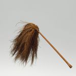 A fly-flap or whisk, made of coconut fibres, affixed to a wooden handle of reddish–brown colour, polished and bulged at both ends and a tassel of blackish-brown coconut fibres.