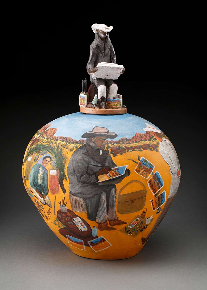 A bulbous pot painted in bright colours, showing a view of an Aboriginal artists at work in the desert. The pot's lid is a sculpted figure of a seated artist.