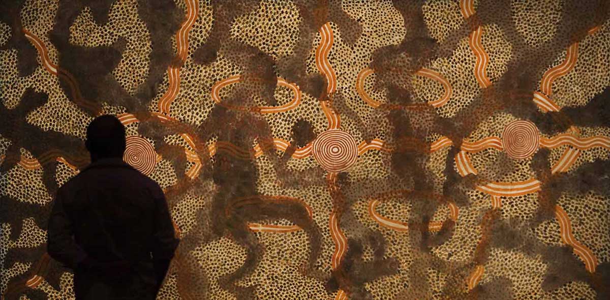 silhouetted man standing in front of painting featuring three concentric circles, centre bisected by shadowy, wavy crossed lines and figures. Others surrounded by oval shape, four wavy lines in diagonal crosses from ovals. Four other ovals around centre circles. White ground with patches of black and yellow dots, and red dots on black
