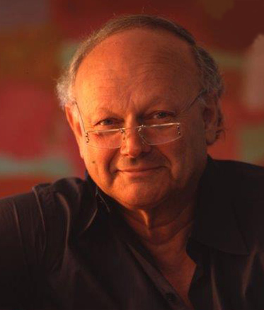 Colour photo portrait of a balding, middle-aged man wearing reading glasses. He is smiling at the camera, and wearing a dark shirt. It appears to have been taken in his studio.