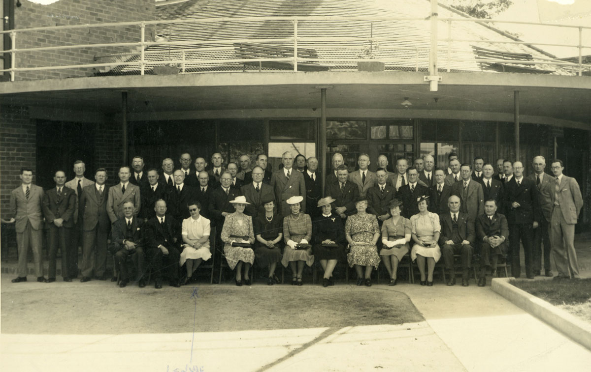 Posed photo of about 20 men standing behind six women sitting.