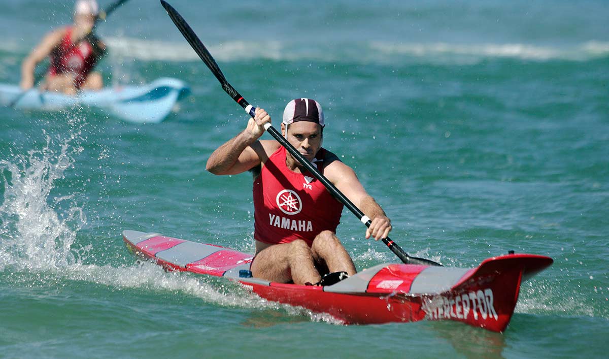 A man in red singlet with Yamaha printed on it paddles a surf ski. Another competitor is visible behind him.