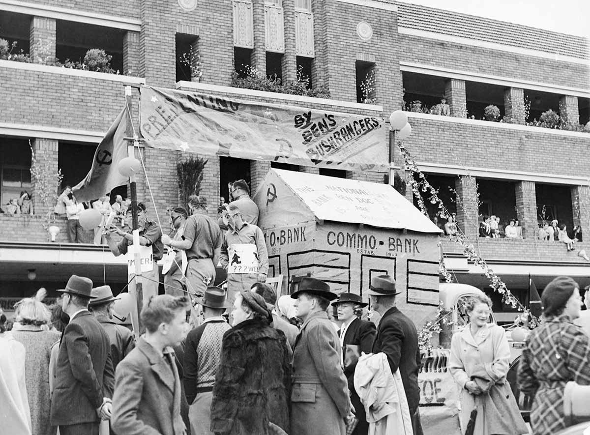 A float with a mocked-up building with the words on ‘Commo Bank’ painted on the side. It is partly obscured by crowds in the street.